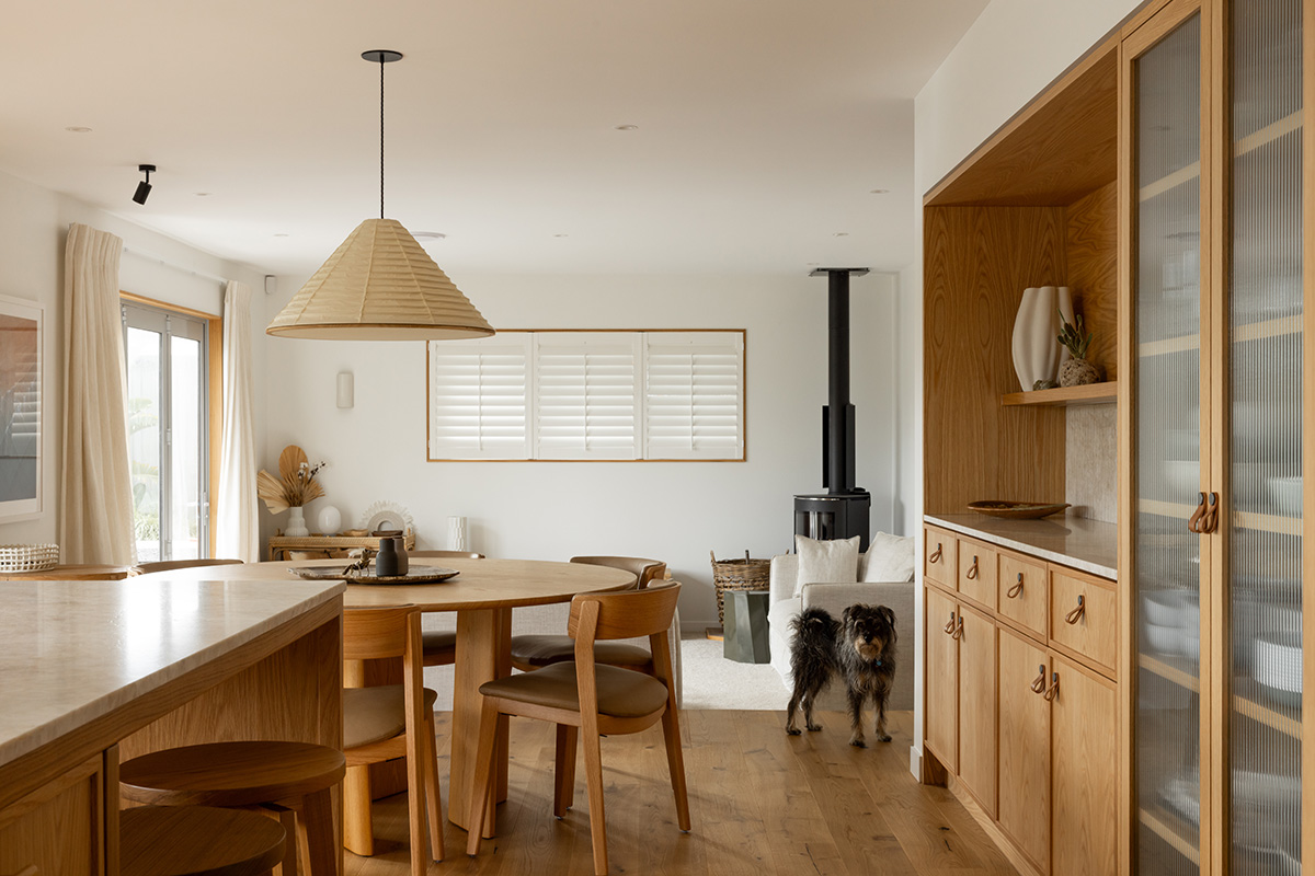 Open-plan dining space in a coastal home with round table, light timber chairs, pale stone island and fluted glass cabinetry.