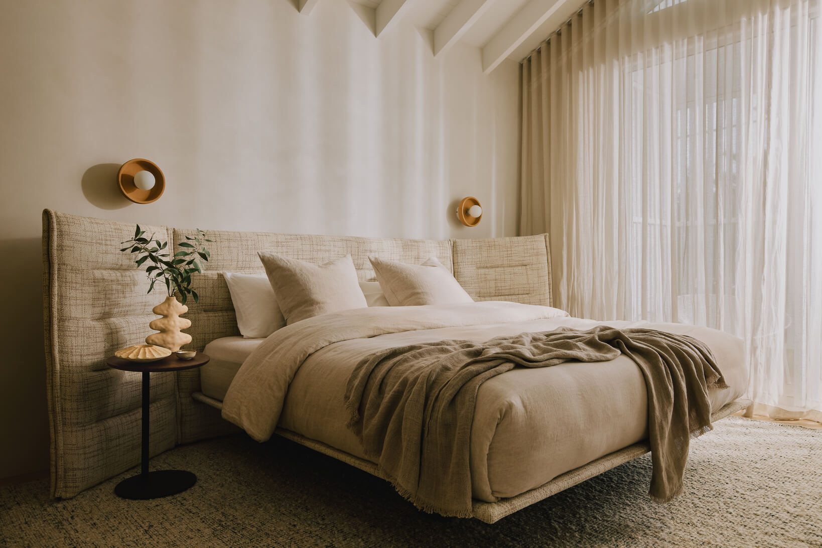 A king size bed with channelled headboard and extended side panels, styled with neutral bedding and round timber side table.